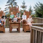 Acht Kinder sitzen auf großen Holzstühlen auf einer Außenterrasse, umgeben von Holzgeländern und Bäumen im Hintergrund.