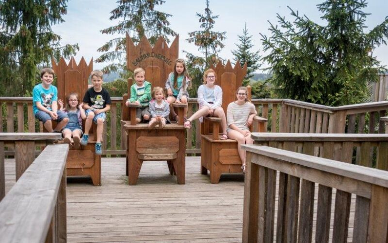 Acht Kinder sitzen auf großen Holzstühlen auf einer Außenterrasse, umgeben von Holzgeländern und Bäumen im Hintergrund.