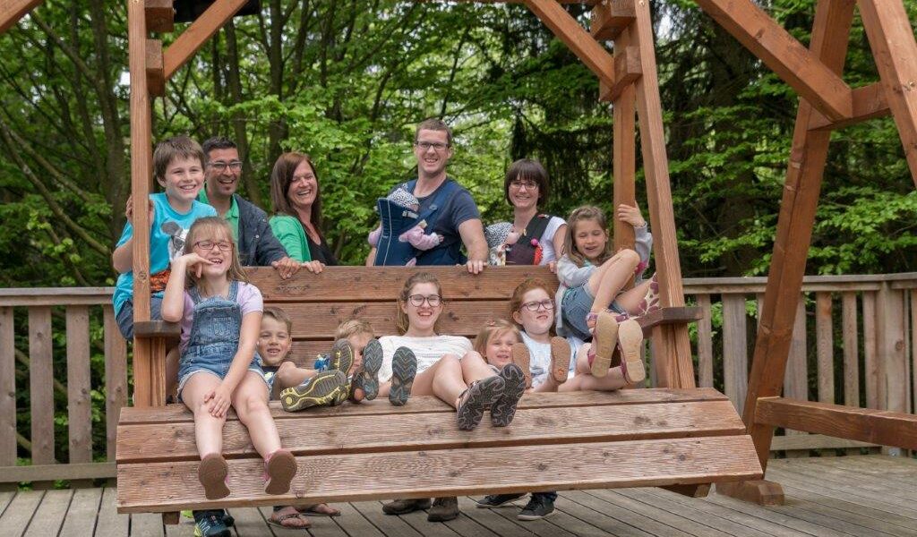 Eine Gruppe von Erwachsenen und Kindern sitzt und steht auf einer großen Holzschaukel auf einem Deck, im Hintergrund sind Bäume zu sehen.