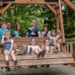 Eine Gruppe von Erwachsenen und Kindern sitzt und steht auf einer großen Holzschaukel auf einem Deck, im Hintergrund sind Bäume zu sehen.