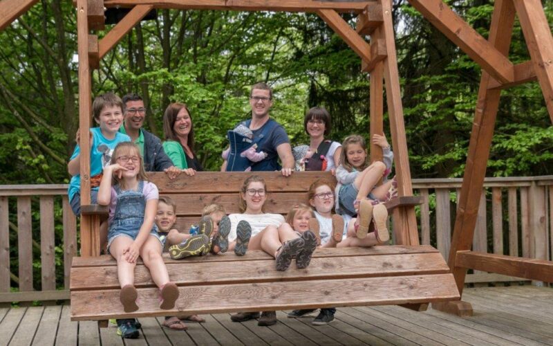 Eine Gruppe von Erwachsenen und Kindern sitzt und steht auf einer großen Holzschaukel auf einem Deck, im Hintergrund sind Bäume zu sehen.