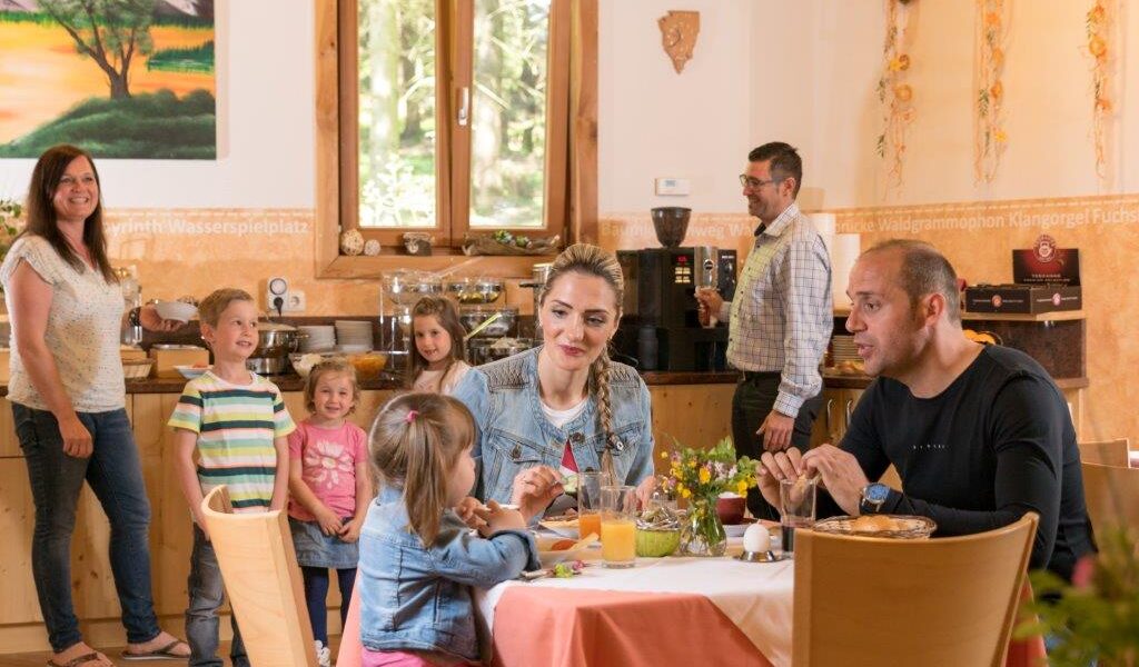 Eine Familie mit Kindern sitzt an einem Tisch und frühstückt in einem hellen Speisesaal, während eine andere Gruppe im Hintergrund an einem Buffet steht.