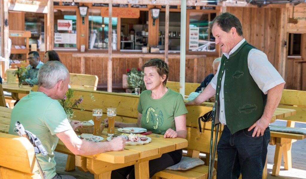 Zwei Personen sitzen an einem Holztisch im Freien mit Essen und Bier und unterhalten sich mit einem stehenden Mann in traditioneller Kleidung in der Nähe eines rustikalen Holzgebäudes.