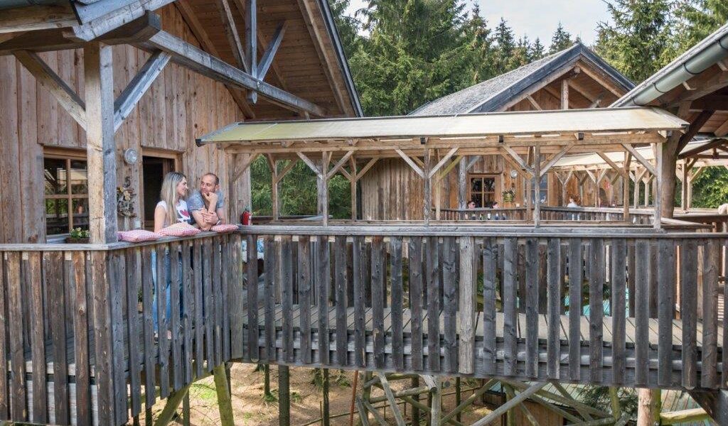 Zwei Personen stehen auf dem Balkon einer Holzhütte, die durch einen überdachten Gang verbunden und von Bäumen umgeben ist.