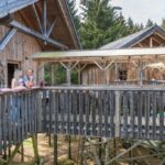 Zwei Personen stehen auf dem Balkon einer Holzhütte, die durch einen überdachten Gang verbunden und von Bäumen umgeben ist.