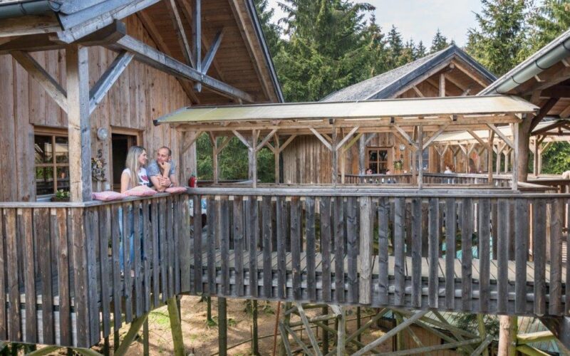 Zwei Personen stehen auf dem Balkon einer Holzhütte, die durch einen überdachten Gang verbunden und von Bäumen umgeben ist.