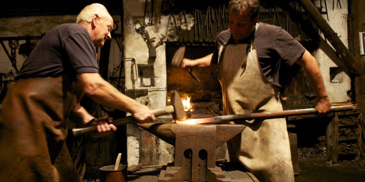 Zwei Schmiede in Schürzen arbeiten zusammen und hämmern eine erhitzte Metallstange auf einen Amboss in einer schwach beleuchteten Werkstatt, in der hinter ihnen Werkzeuge an der Wand hängen.