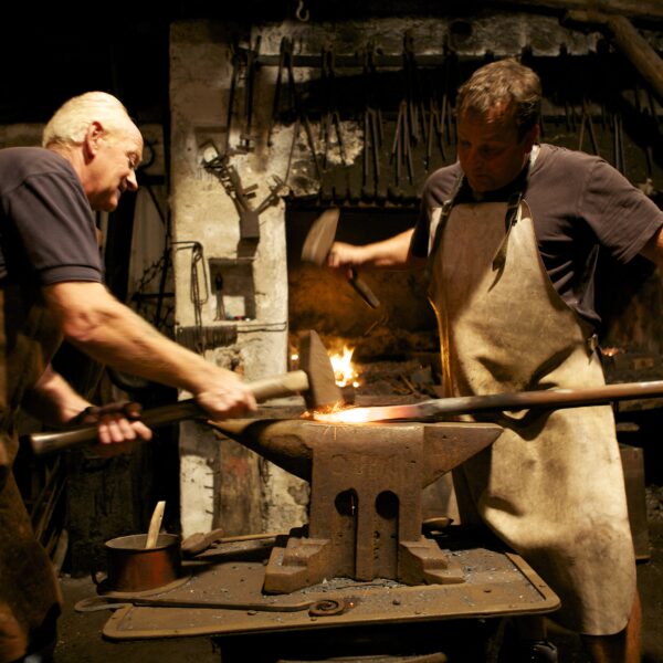 Zwei Schmiede in Schürzen arbeiten zusammen und hämmern eine erhitzte Metallstange auf einen Amboss in einer schwach beleuchteten Werkstatt, in der hinter ihnen Werkzeuge an der Wand hängen.