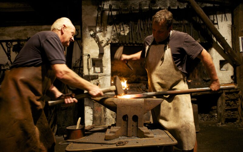 Zwei Schmiede in Schürzen arbeiten zusammen und hämmern eine erhitzte Metallstange auf einen Amboss in einer schwach beleuchteten Werkstatt, in der hinter ihnen Werkzeuge an der Wand hängen.