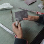 Eine Person glättet mit Schleifpapier und Feilen ein Stück Stein auf einem Tisch, der mit Werkzeugen und einem Steinfragment vollgestopft ist.