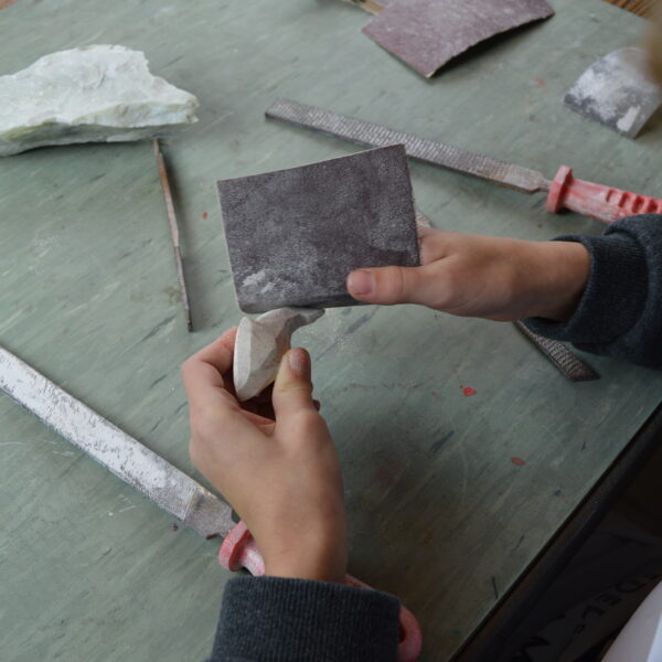 Eine Person glättet mit Schleifpapier und Feilen ein Stück Stein auf einem Tisch, der mit Werkzeugen und einem Steinfragment vollgestopft ist.