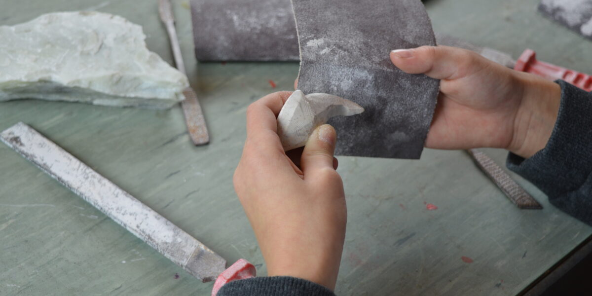 Eine Person glättet mit Schleifpapier eine kleine Steinskulptur auf einem Arbeitstisch mit Feilen und anderen Werkzeugen.