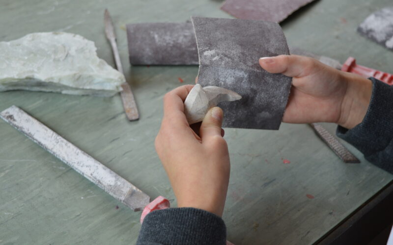Eine Person glättet mit Schleifpapier eine kleine Steinskulptur auf einem Arbeitstisch mit Feilen und anderen Werkzeugen.