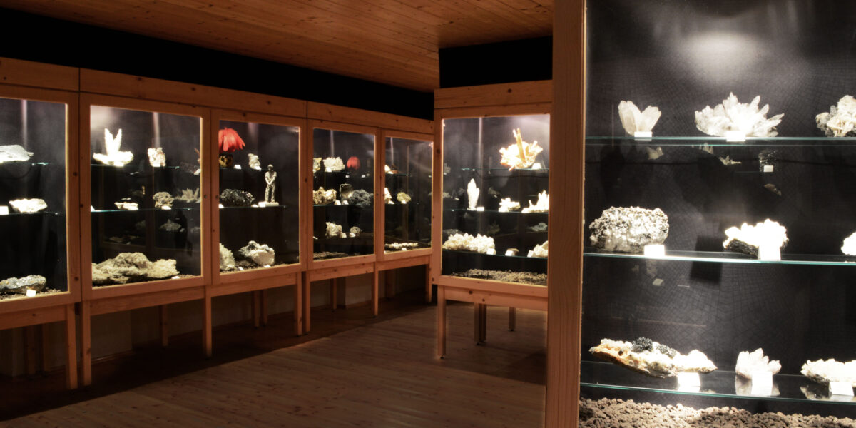 In einem schwach beleuchteten Museumsraum sind verschiedene Mineralien und Kristalle in Vitrinen an Holzwänden und -böden ausgestellt.