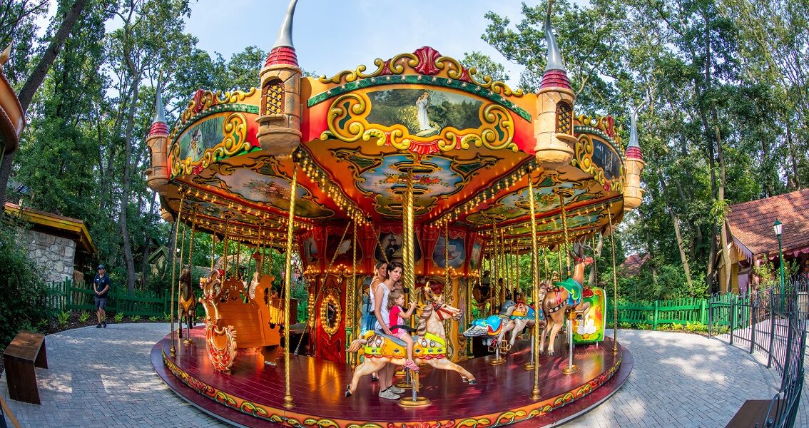 Ein farbenfrohes Karussell mit Kindern und Erwachsenen, die auf verschiedenen Tierfiguren reiten, in einer von Bäumen und Grün umgebenen Parklandschaft.