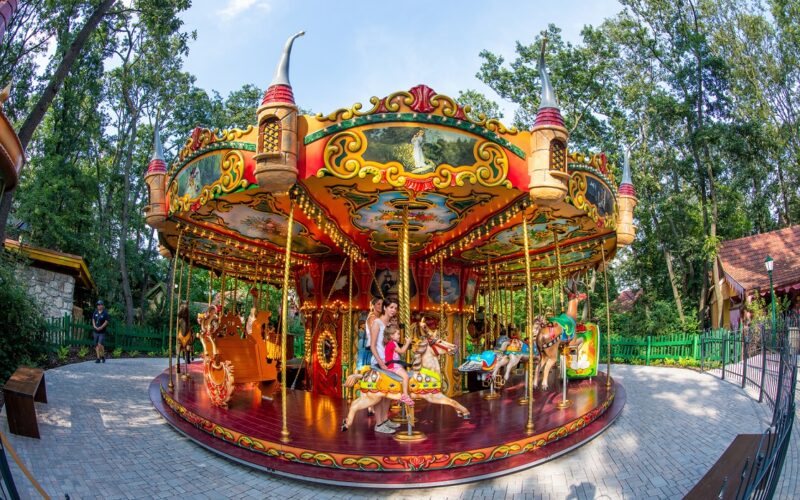 Ein farbenfrohes Karussell mit Kindern und Erwachsenen, die auf verschiedenen Tierfiguren reiten, in einer von Bäumen und Grün umgebenen Parklandschaft.