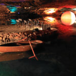 In einer unterirdischen Höhle liegt ein kleines Holzboot vor Anker. Auf einer Plattform sind Stühle und Notenständer angeordnet, und die Felswände werden von bunten Lichtern beleuchtet.