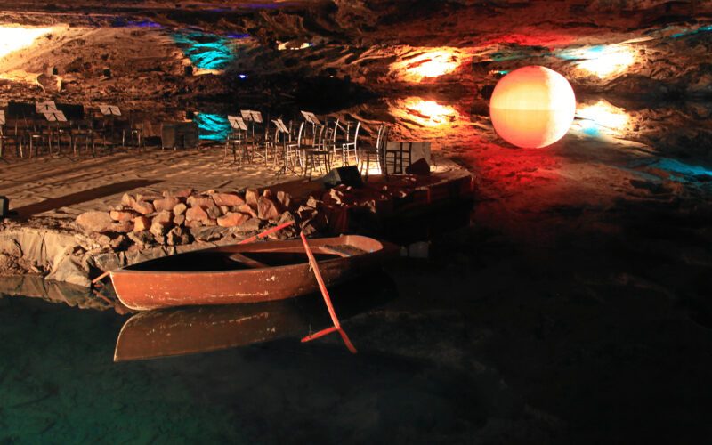 In einer unterirdischen Höhle liegt ein kleines Holzboot vor Anker. Auf einer Plattform sind Stühle und Notenständer angeordnet, und die Felswände werden von bunten Lichtern beleuchtet.