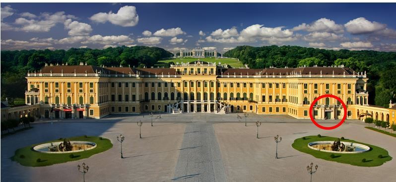 Ein großer gelber Barockpalast mit symmetrischen Flügeln, zwei Springbrunnen davor und einem roten Kreis, der einen Bereich auf der rechten Seite des Gebäudes markiert.