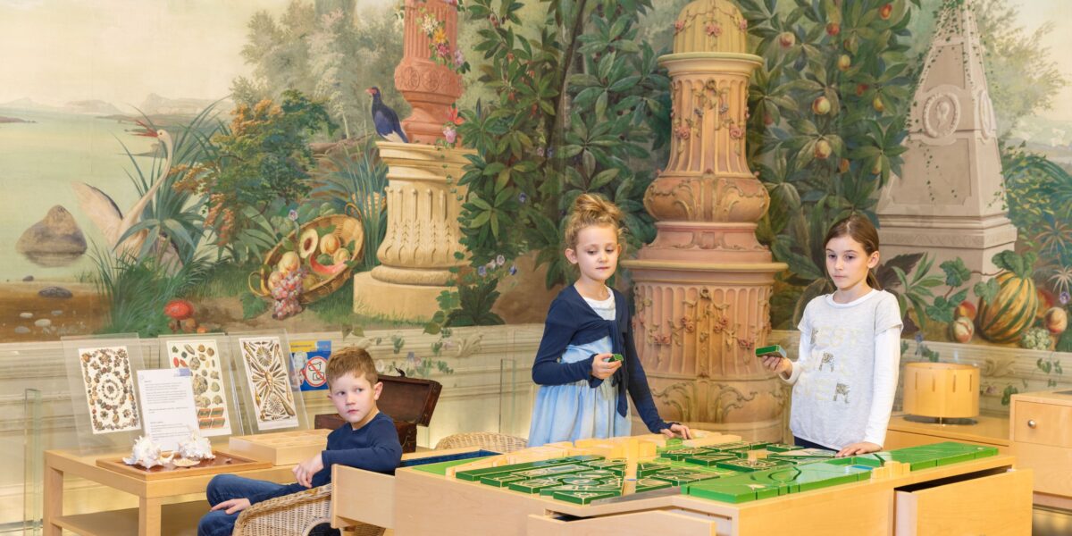 Drei Kinder spielen mit einem Tisch-Labyrinthspiel in einem dekorierten Raum mit einem gemalten Wandgemälde zum Thema Natur und verzierten Säulen im Hintergrund.