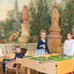 Drei Kinder spielen mit einem Tisch-Labyrinthspiel in einem dekorierten Raum mit einem gemalten Wandgemälde zum Thema Natur und verzierten Säulen im Hintergrund.