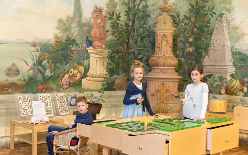 Drei Kinder spielen mit einem Tisch-Labyrinthspiel in einem dekorierten Raum mit einem gemalten Wandgemälde zum Thema Natur und verzierten Säulen im Hintergrund.