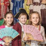 Drei Kinder in historischen Kostümen posieren mit Spitzenfächern vor einer Ausstellung mit alten Kleidern.