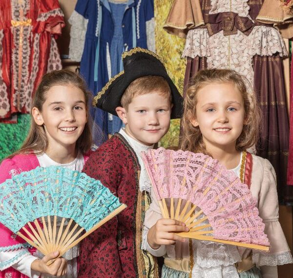 Drei Kinder in historischen Kostümen posieren mit Spitzenfächern vor einer Ausstellung mit alten Kleidern.