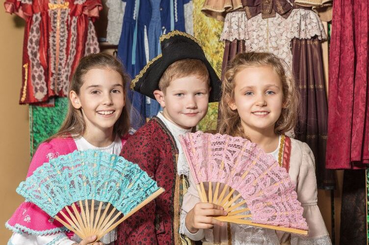 Drei Kinder in historischen Kostümen posieren mit Spitzenfächern vor einer Ausstellung mit alten Kleidern.