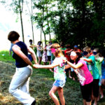 Eine Gruppe von Kindern, einige mit verbundenen Augen, folgt einem Erwachsenen in einer Reihe durch eine von Bäumen umgebene Wiese.