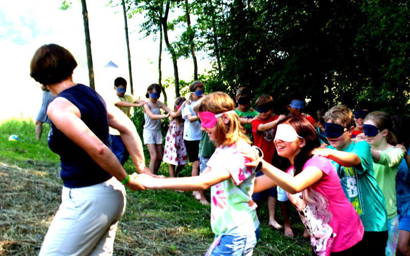 Eine Gruppe von Kindern, einige mit verbundenen Augen, folgt einem Erwachsenen in einer Reihe durch eine von Bäumen umgebene Wiese.