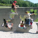 Kinder klettern und spielen auf einer Betonwand mit unregelmäßig geformten Löchern auf einem Spielplatz im Freien.