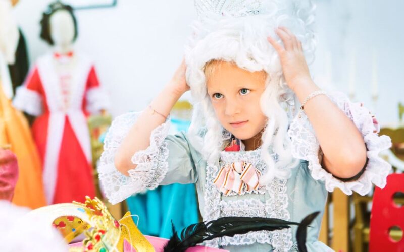 Ein kleines Kind mit weißer Perücke, Krone und historischem Kostüm richtet seine Perücke, umgeben von farbenfrohen Kostümaccessoires.