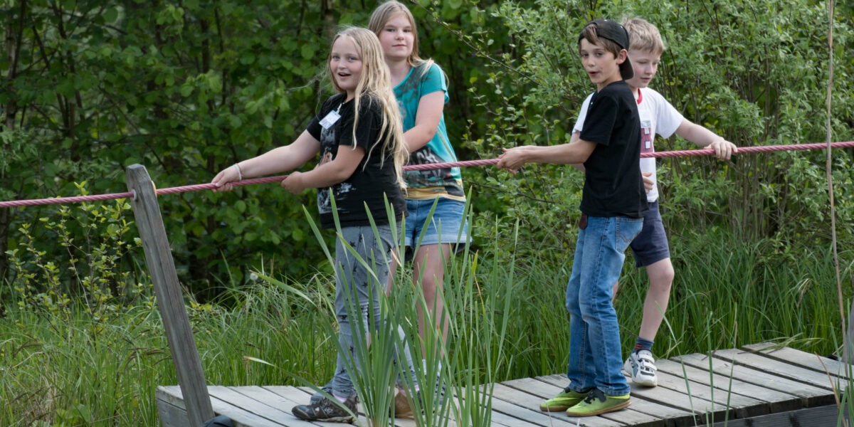Vier Kinder stehen auf einem Holzfloß und ziehen sich an einem Seil über ein Gewässer, das von Grün umgeben ist.