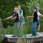 Vier Kinder stehen auf einem Holzfloß und ziehen sich an einem Seil über ein Gewässer, das von Grün umgeben ist.