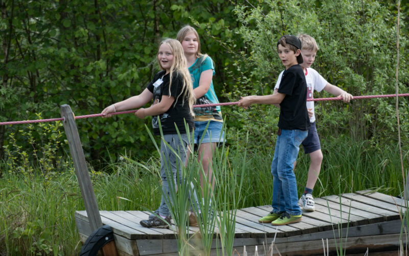 Vier Kinder stehen auf einem Holzfloß und ziehen sich an einem Seil über ein Gewässer, das von Grün umgeben ist.