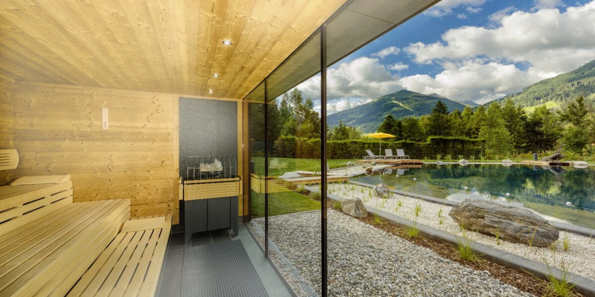 Die holzgetäfelte Sauna mit großer Glaswand bietet einen Blick auf einen natürlichen Schwimmteich, einen Garten und eine malerische Berglandschaft unter wolkenverhangenem Himmel.