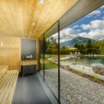 Die holzgetäfelte Sauna mit großer Glaswand bietet einen Blick auf einen natürlichen Schwimmteich, einen Garten und eine malerische Berglandschaft unter wolkenverhangenem Himmel.