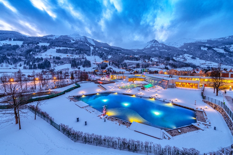 In der Abenddämmerung beleuchteter Freibadkomplex, umgeben von schneebedecktem Boden, mit Bergen und Gebäuden im Hintergrund unter einem bewölkten Himmel.