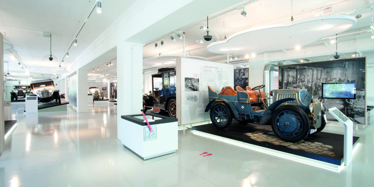 Eine moderne Museumsausstellung zeigt Oldtimer hinter Glas, mit Informationstafeln und heller Deckenbeleuchtung in einem sauberen, weißen Innenraum.