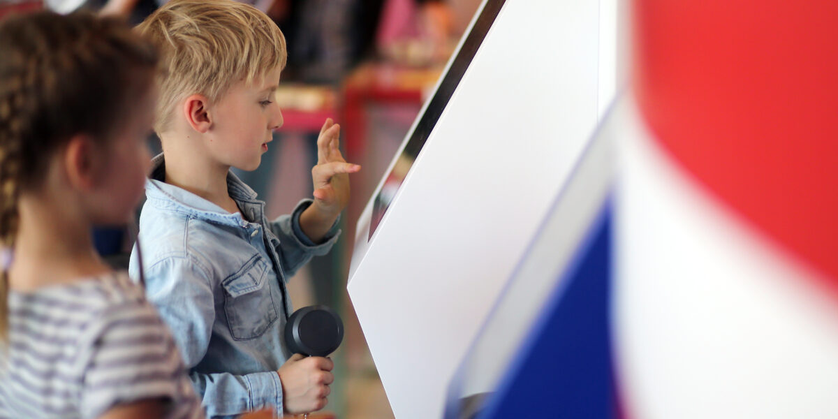 Zwei Kinder interagieren mit einem Touchscreen-Kiosk; der Junge berührt den Bildschirm und hält einen telefonähnlichen Hörer in der Hand, während das Mädchen neben ihm steht.