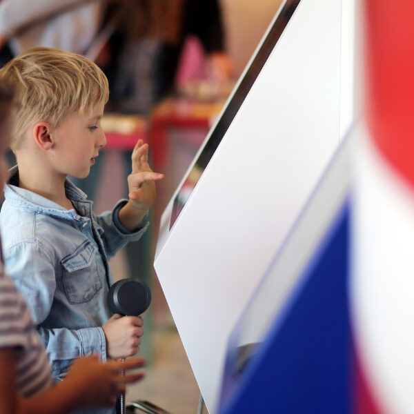 Zwei Kinder interagieren mit einem Touchscreen-Kiosk; der Junge berührt den Bildschirm und hält einen telefonähnlichen Hörer in der Hand, während das Mädchen neben ihm steht.