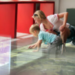 Eine Frau und ein kleiner Junge knien auf dem Boden und untersuchen eine Karte oder einen Schaukasten unter Glas in einem hell erleuchteten Raum mit roten Ständern.