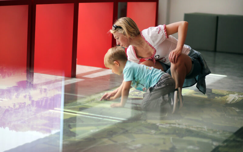 Eine Frau und ein kleiner Junge knien auf dem Boden und untersuchen eine Karte oder einen Schaukasten unter Glas in einem hell erleuchteten Raum mit roten Ständern.