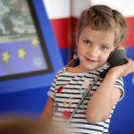 Ein junges Mädchen in einem gestreiften Hemd hält sich einen Telefonhörer ans Ohr und steht neben einem Display mit gelben Sternen auf blauem Hintergrund.
