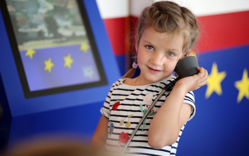 Ein junges Mädchen in einem gestreiften Hemd hält sich einen Telefonhörer ans Ohr und steht neben einem Display mit gelben Sternen auf blauem Hintergrund.