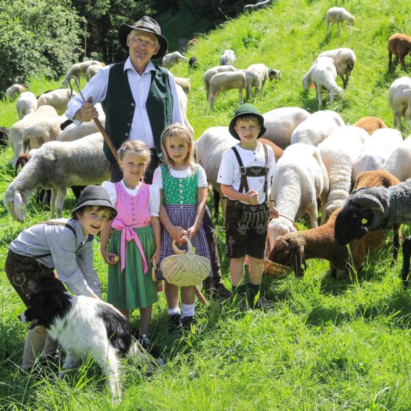 Ein Mann und vier Kinder in traditioneller Kleidung stehen auf einem grasbewachsenen Feld mit einer Schafherde und einem schwarz-weißen Hund.