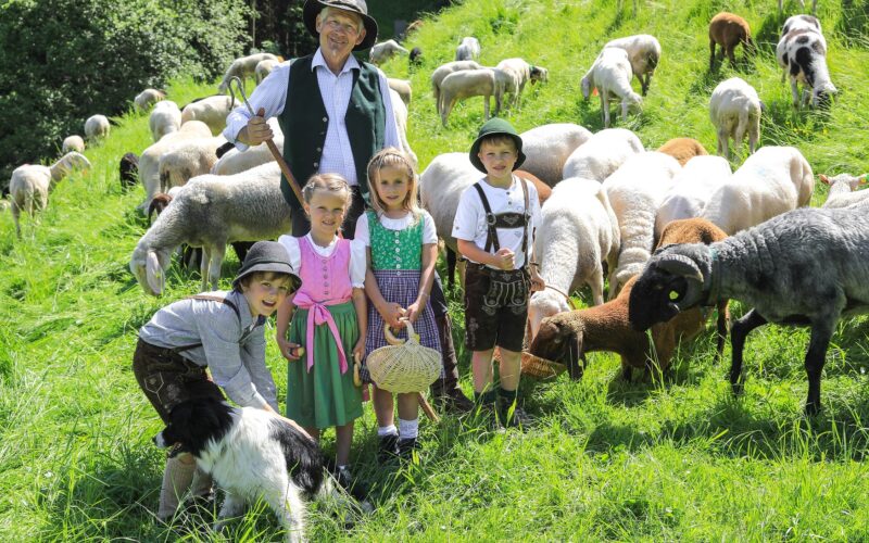 Ein Mann und vier Kinder in traditioneller Kleidung stehen auf einem grasbewachsenen Feld mit einer Schafherde und einem schwarz-weißen Hund.