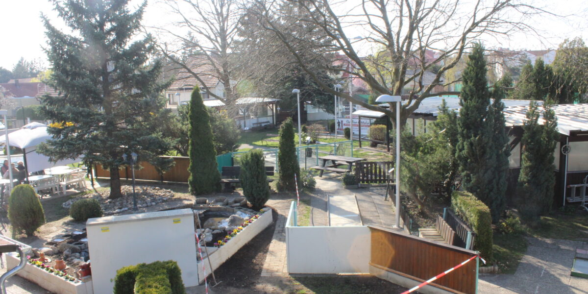 Naherholungsgebiet mit Bäumen, Wegen und kleinen Gebäuden, das an einem sonnigen Tag teilweise mit rot-weißem Band abgesperrt ist.