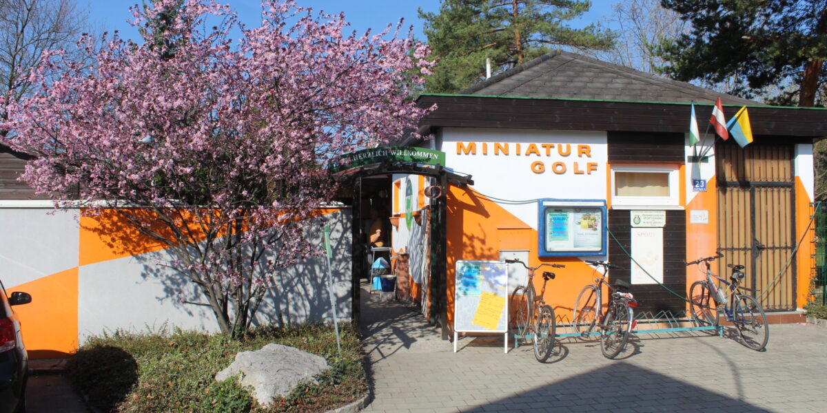 Ein kleines Gebäude mit der Aufschrift "Miniatur Golf", vor dem Fahrräder geparkt sind, ein blühender Baum zur Linken und orange und weiß gestrichene Wände unter einem klaren Himmel.